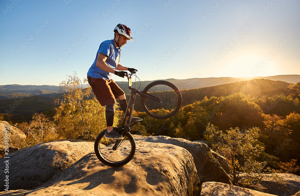 Athletic cyclist balancing on back wheel on trial bike. Sportsman biker making acrobatic trick on the edge of big boulder on the top of mountain at sunset. Concept of extreme sport active lifestyle Athletic cyclist balancing on back wheel on trial bike. Sportsman biker making acrobatic trick on the edge of big boulder on the top of mountain at sunset. Concept of extreme sport active lifestyle