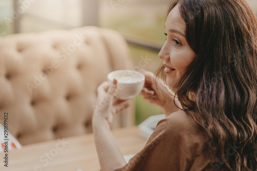 Charming brunette woman with long curly hair sitting at window in cafe with cup of coffee in hands