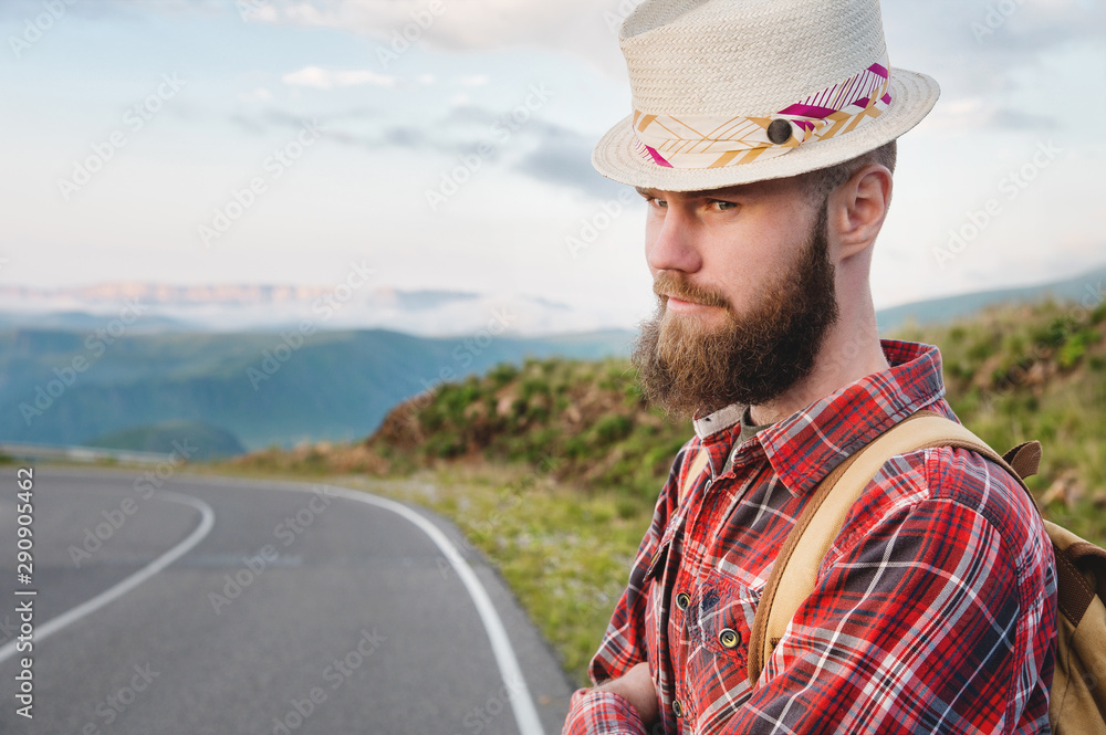 Portrait of a bearded happy serious traveler hipster with a backpack in a plaid shirt and a hat next to an unknown car stands on the road at sunset in the mountains. Happy and confident travel concept Portrait of a bearded happy serious traveler hipster with a backpack in a plaid shirt and a hat next to an unknown car stands on the road at sunset in the mountains. Happy and confident travel concept