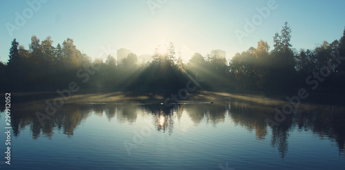 Panorama of a lake with fog and bright sunbeams passing through the crowns of trees