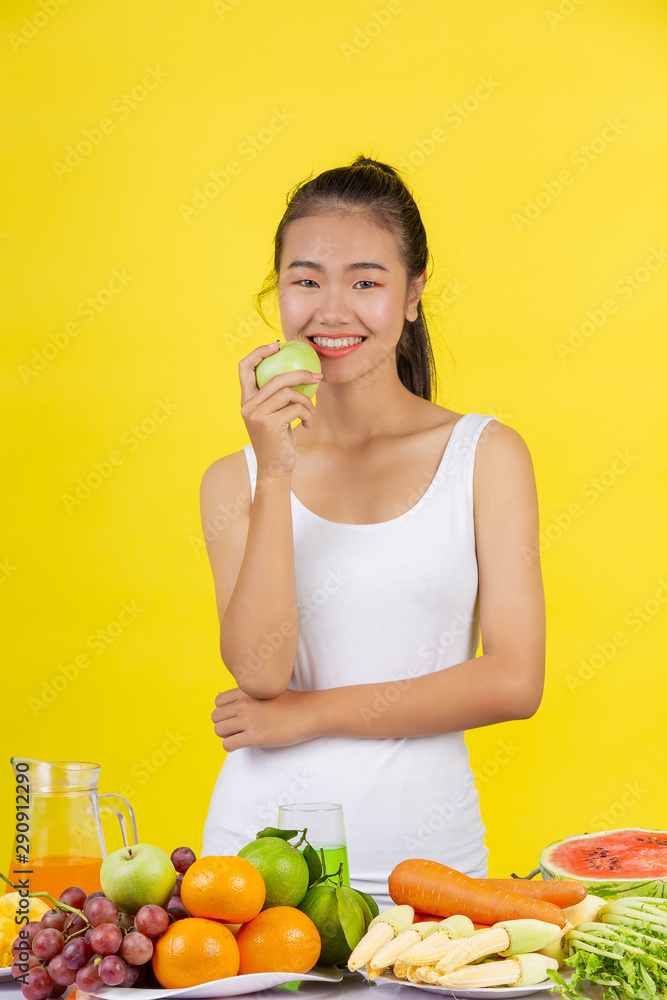 An Asian woman holding a green apple with her right hand, and on the table there are many fruits. An Asian woman holding a green apple with her right hand, and on the table there are many fruits.