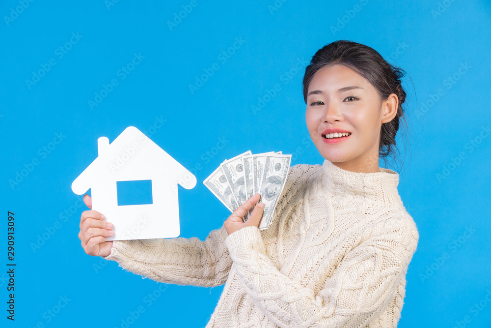 A beautiful woman wearing a new long-sleeved white carpet that holds the house symbol and dollar bills on a blue background. Trading concept. A beautiful woman wearing a new long-sleeved white carpet that holds the house symbol and dollar bills on a blue background. Trading concept.