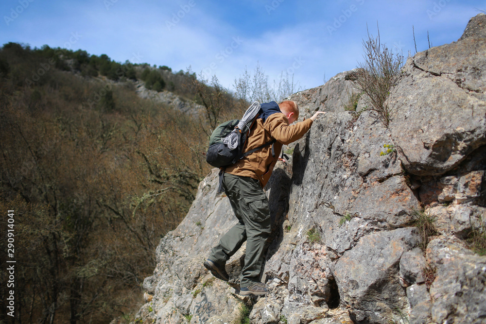 Red-haired man in a brown windbreaker with a backpack and a rope climbs a cliff in the spring. In the background is a forest without foliage.