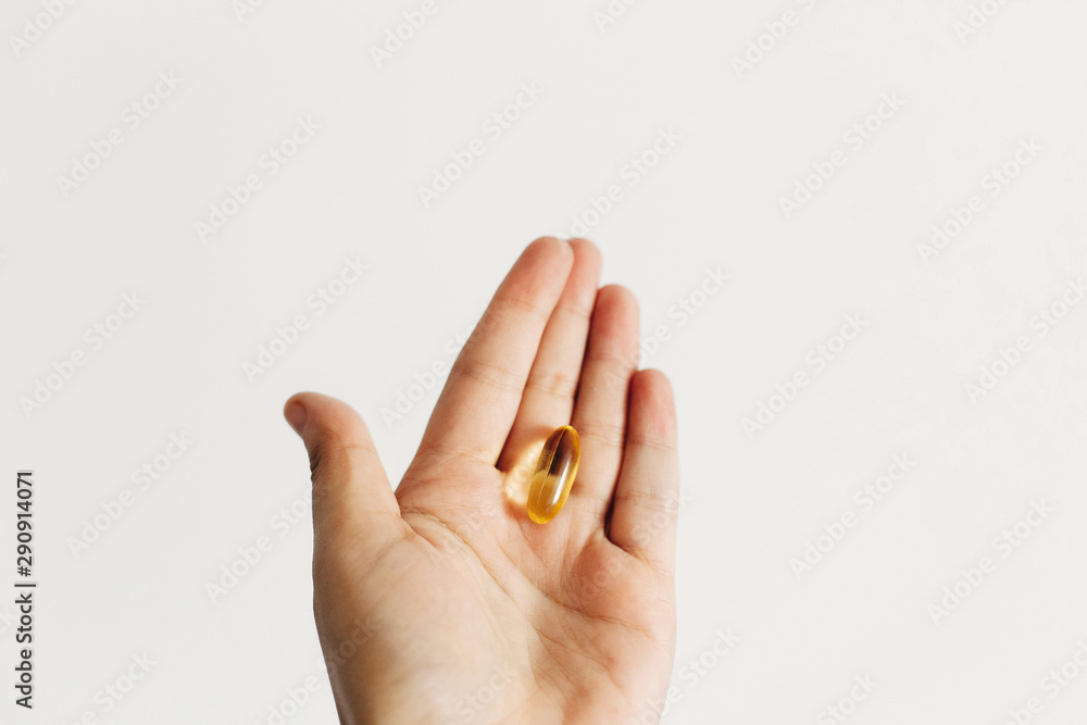 Hand holding omega 3 capsule on white wall background. Morning dose of vitamin. Fish oil in pills. Dietary supplements. Health support and treatment. Biologically active additives Hand holding omega 3 capsule on white wall background. Morning dose of vitamin. Fish oil in pills. Dietary supplements. Health support and treatment. Biologically active additives
