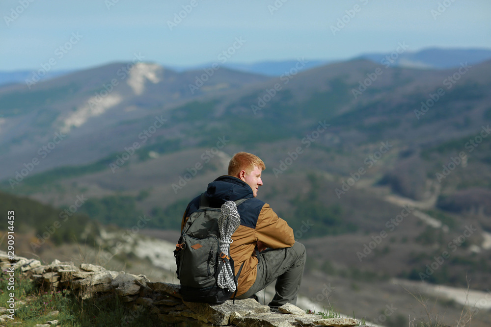 Red-haired man in a brown-and-blue windbreaker with green backpack sits on the slope of Mangup plateau in Crimea. Blurred spring forest is visible in valley in the background.