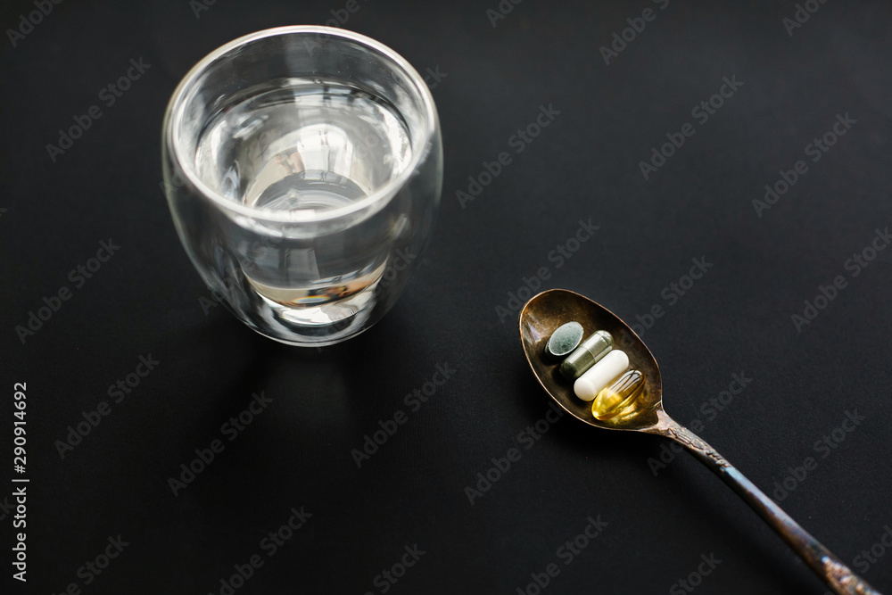 Dietary supplements. Dose of daily vitamins. Omega 3, spirulina, chlorophyll, magnesium capsules on spoon and glass of water on black dark background. Health support and treatment