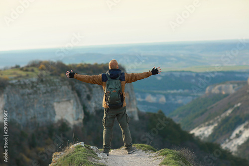 A red-haired man in a brown and blue windbreaker with a backpack stands with open arms on the edge of a cliff on Mangup plateau. Crimean mountains are visible in the background.