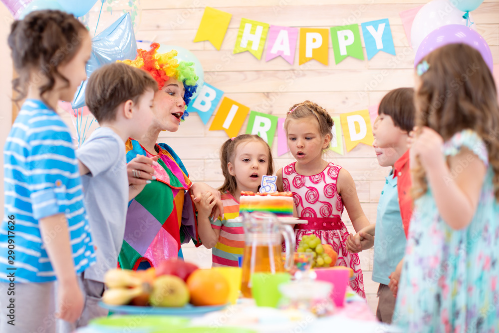 Preschool children holding by hands and blowing candle up. Little kid girl 5 years celebrating her birthday together with friends and clown Preschool children holding by hands and blowing candle up. Little kid girl 5 years celebrating her birthday together with friends and clown