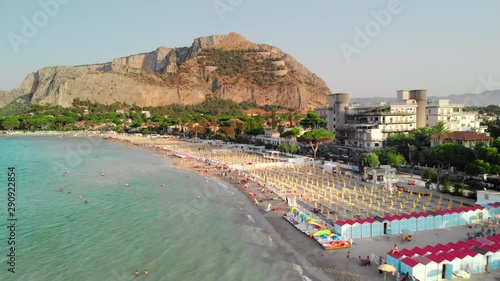 Palermo, Sicily, Italy: The camera slides over ocean coastline. Many vacationers and tourists on the beach. Symmetrically arranged parasols from the sun. Big rock on background. Aerial drone shot