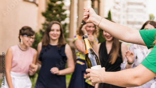 Young woman opening a bottle of champagne with female friends standing in background laughing and having fun. Young people celebrating with champagne at party outdoors.