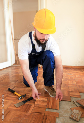 Construction worker and constructor is working on renovation of apartment. Handyman is removing old wooden parquet flooring using yellow hammer and scraping tool. Builder dismantles parquet floor.