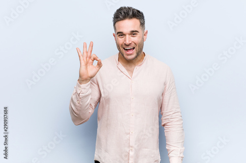 Young handsome cool man winks an eye and holds an okay gesture with hand.
