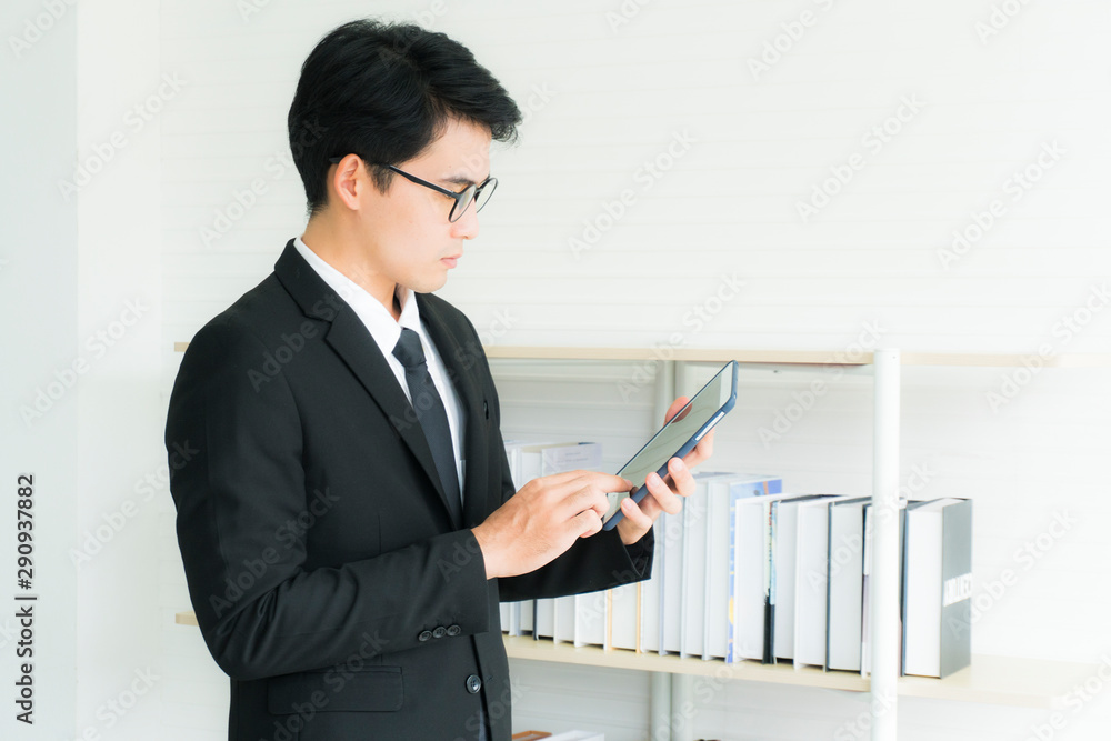 Young Asian handsome businessman wearing black suit holding tablet standing near the bookshelf in his office Young Asian handsome businessman wearing black suit holding tablet standing near the bookshelf in his office