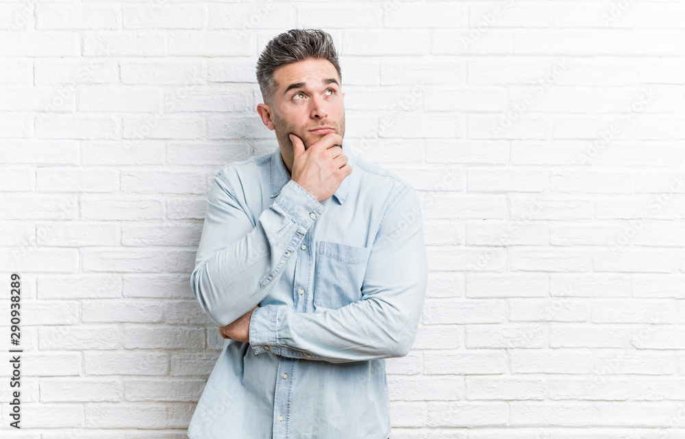 Young handsome man against a bricks wall looking sideways with doubtful and skeptical expression.