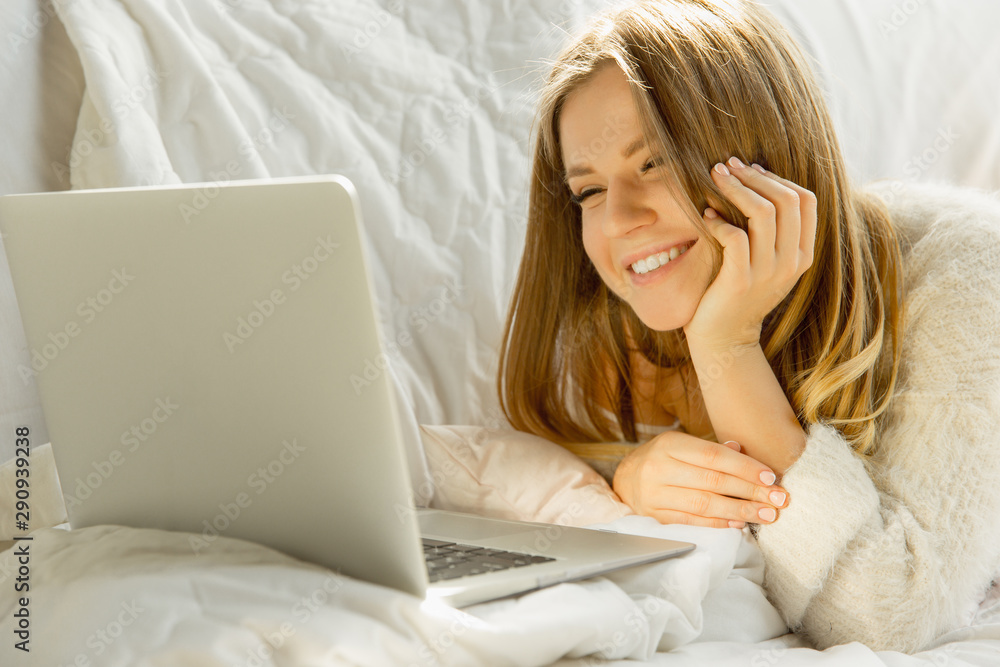 Relaxing at home. Beautiful young woman using her laptop while lying on sofa at home with warm sunlight. Caucasian blonde female model has weekend time for resting. Watches cinema or makes vlog. Relaxing at home. Beautiful young woman using her laptop while lying on sofa at home with warm sunlight. Caucasian blonde female model has weekend time for resting. Watches cinema or makes vlog.