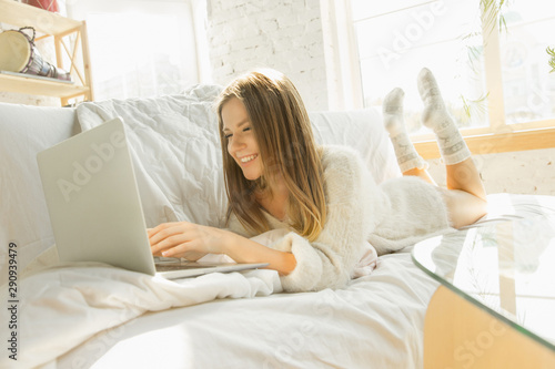 Relaxing at home. Beautiful young woman using her laptop while lying on sofa at home with warm sunlight. Caucasian blonde female model has weekend time for resting. Watches cinema or makes vlog.