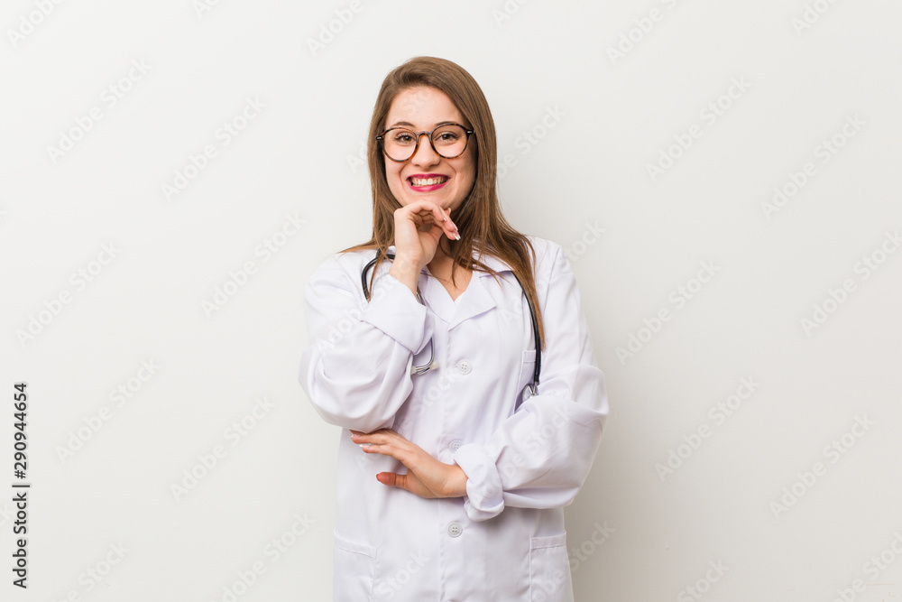 Young doctor woman against a white wall smiling happy and confident, touching chin with hand. Young doctor woman against a white wall smiling happy and confident, touching chin with hand.