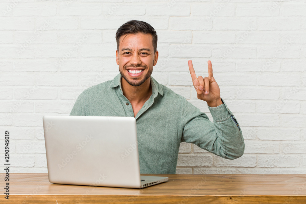 Young filipino man sitting working with his laptop showing a horns gesture as a revolution concept. Young filipino man sitting working with his laptop showing a horns gesture as a revolution concept.