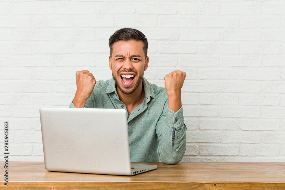 Young filipino man sitting working with his laptop cheering carefree and excited. Victory concept. Young filipino man sitting working with his laptop cheering carefree and excited. Victory concept.