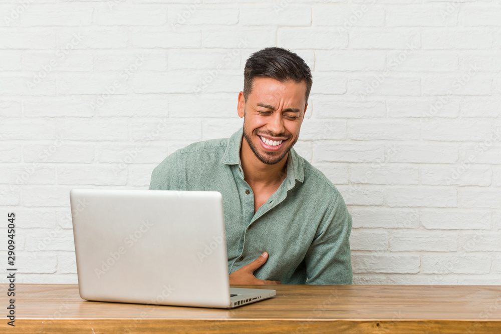 Young filipino man sitting working with his laptop laughs happily and has fun keeping hands on stomach. Young filipino man sitting working with his laptop laughs happily and has fun keeping hands on stomach.