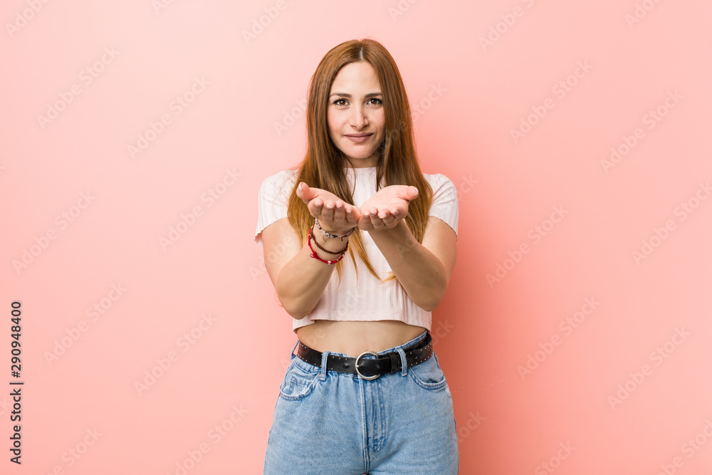 Young redhead ginger woman against a pink wall holding something with palms, offering to camera. Young redhead ginger woman against a pink wall holding something with palms, offering to camera.