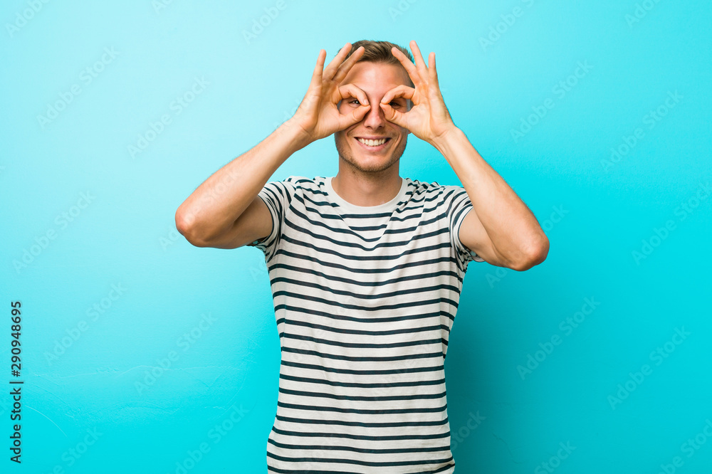 Young caucasian man against a blue wall showing okay sign over eyes Young caucasian man against a blue wall showing okay sign over eyes