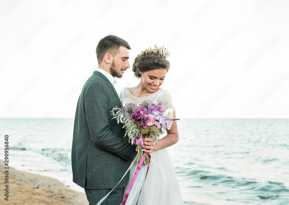 Happy newlyweds stand holding hands on the background of the blue sea. Wedding walk on a sand beach. In the background, blue sky Happy newlyweds stand holding hands on the background of the blue sea. Wedding walk on a sand beach. In the background, blue sky