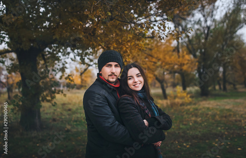 Young caucasian guy with mustache and beard and girl are standing embracing on the background of autumn nature. A pair of lovers dressed in warm clothes, a coat, a scarf, a hat. © shchus