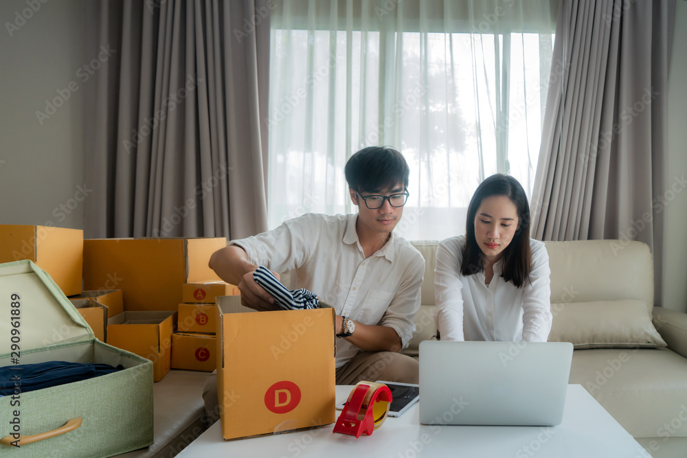 Asian young couple are selling on-line via a computer and helping to check the box in order to send mail in living room at house. Small business startup SME entrepreneur or freelance concept Asian young couple are selling on-line via a computer and helping to check the box in order to send mail in living room at house. Small business startup SME entrepreneur or freelance concept