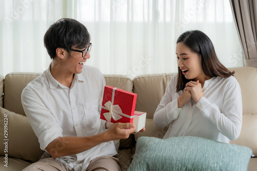 Asian man give a woman a red gift box. She look at the gift in the box and surprise at the wedding anniversary or her birthday in living room at house. Love, relationship, wedding, or relaxing casual