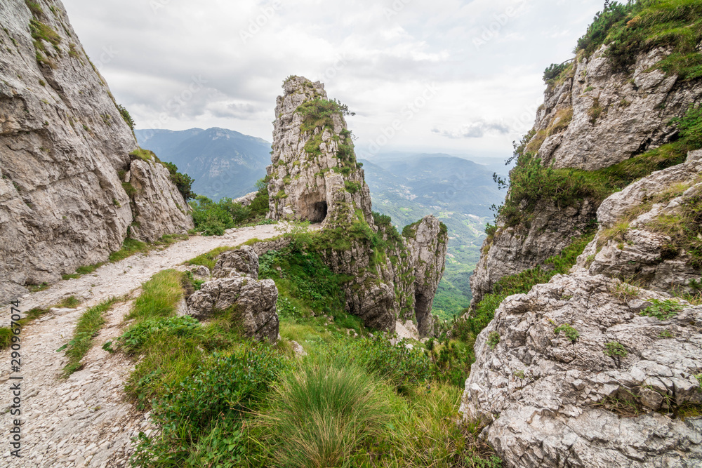 Wide angle shot of an italian mountain landscape, with a trail snaking among rocks in the foreground, and the entrance to a gallery carved into the rock in the background