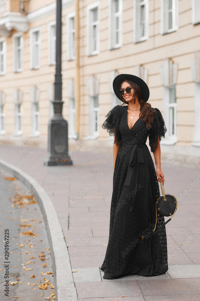 Fototapeta premium Elegant woman in long black dress, hat and sunglasses holding handbag standing and posing at city street. Stylish fashionable warm autumn look. Full length outdoor portrait