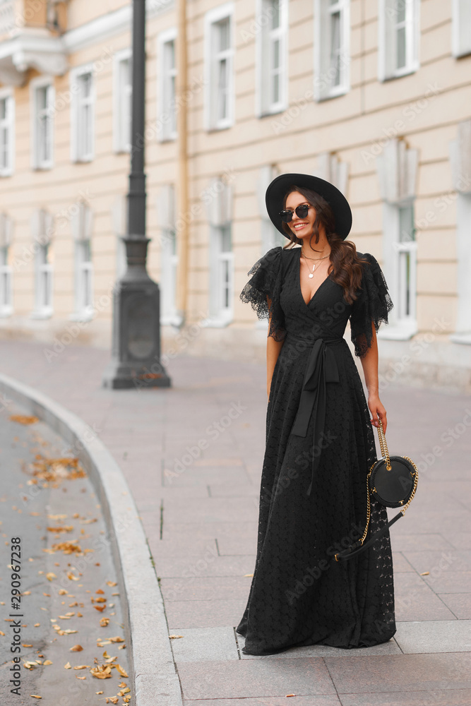 Elegant woman in long black dress, hat and sunglasses holding handbag standing and posing at city street. Stylish fashionable warm autumn look. Full length outdoor portrait