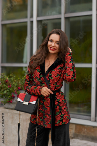 Body length outdoor portrait of young beautiful caucasian woman with long wavy brunette hair in red coat, black trousers and sunglasses with leather handbag posing againsy modern glass and metal wall photo