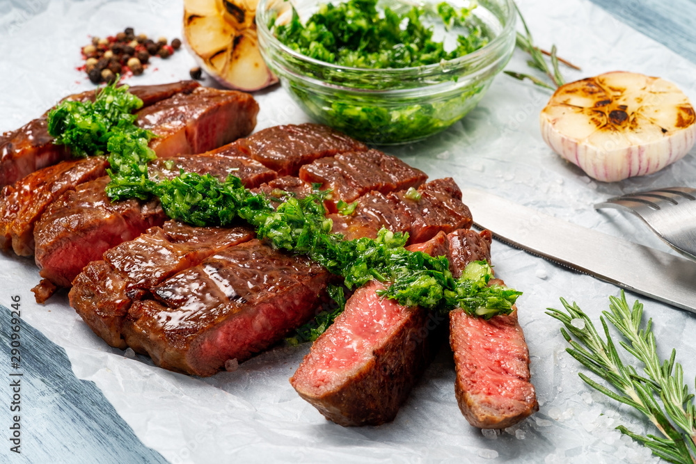 Sliced grilled rib eye beef steak with chimichurri sauce on white paper on wooden background. Concept of cooked steak