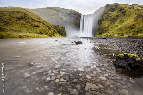 Iconic icelandic landscape with a stream flowing over pebbles in foreground and the Skogafoss waterfall, surrounded by green hills, in the background