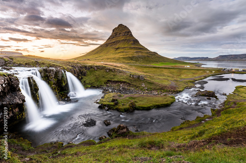 Iconic icelandic landscape at sunset, with a waterfall in the foreground and a conic mountain in the background, under a colorful cloudy sky