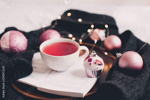 Fresh hot morning cup of tea on open book with Christmas decorations on knitted sweater over Christmas glowing lights at background close up. Good morning. Winter holiday season.