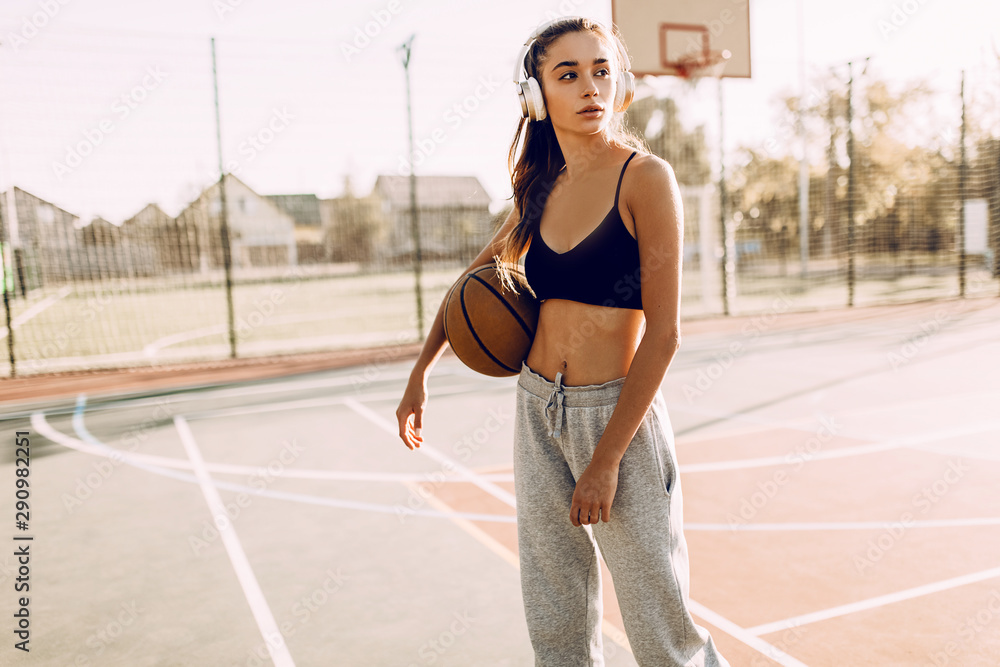 Portrait of a beautiful strong athletic woman outdoors posing while listening to music with headphones holding a basketball.