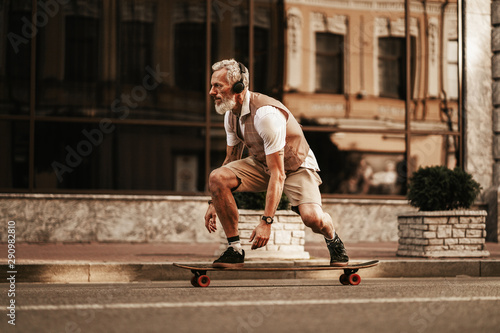 Portrait of bearded hipster man in white shirt on city street. Stylish happy model ride on longboard near road on buildings background