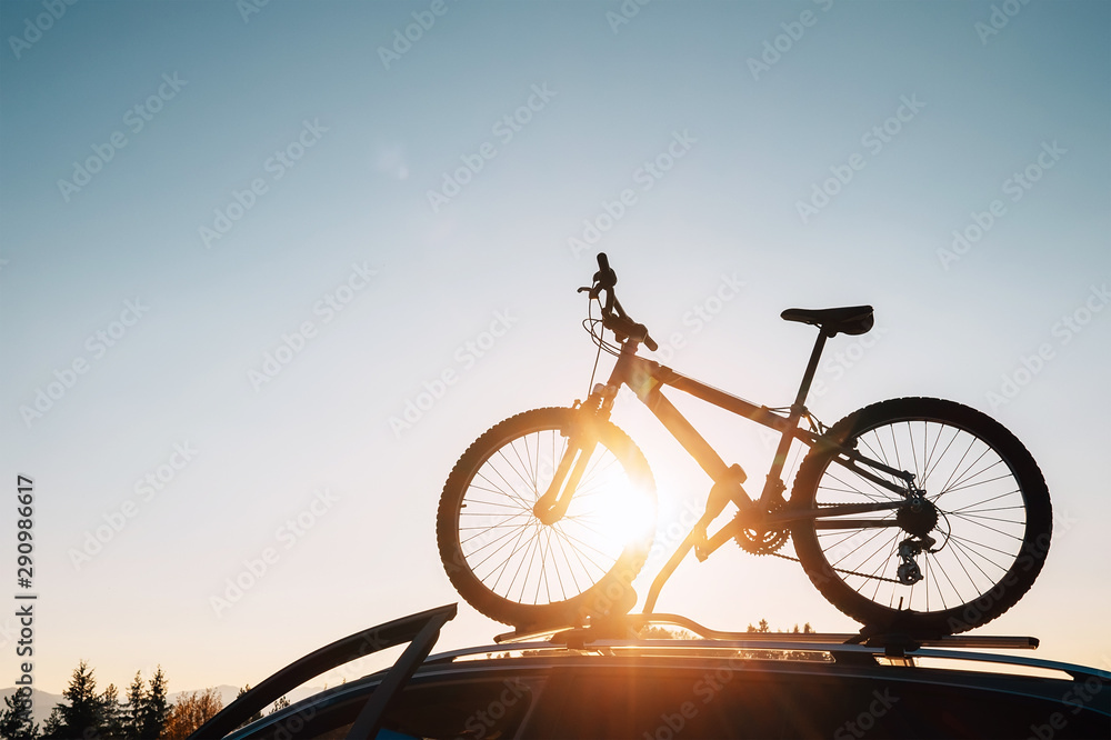 Obraz premium Mounted mountain bicycle silhouette on the car roof with evening sun light rays background. Safe sport items transportation using a car concept image.