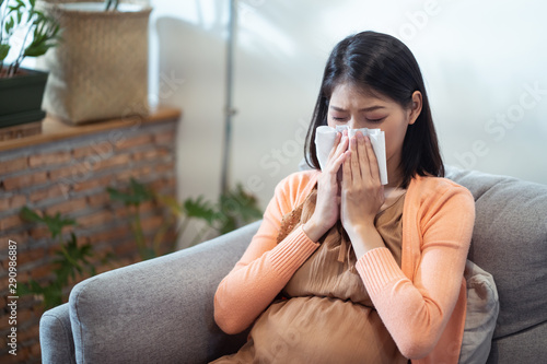 Asian pregnancy woman having a cough and cold fever sitting on sofa at home. She sneezing on tissue paper with eyes closing. Healthcare and medical concern for pregnant concept.