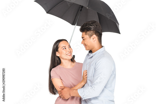happy asian couple hugging and looking at each other while standing under umbrella isolated on white