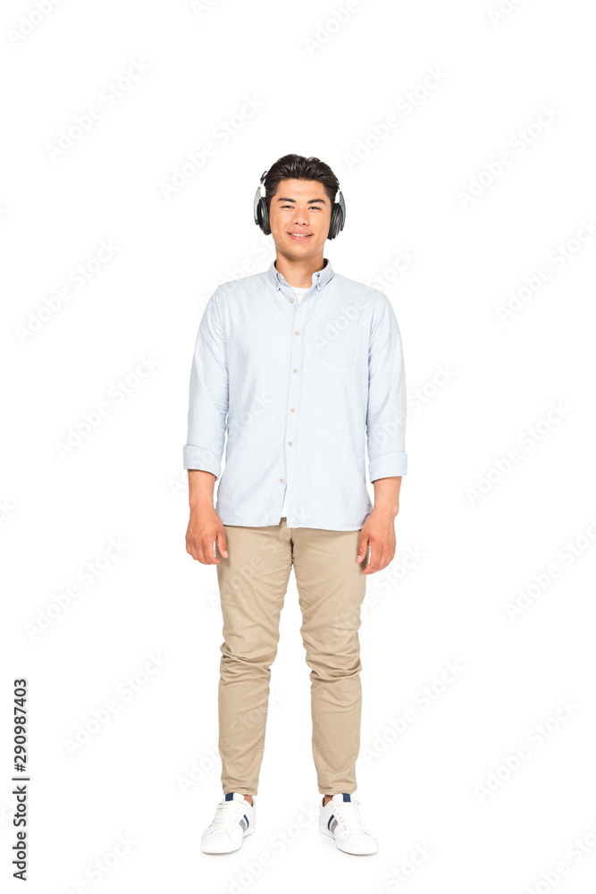 Fototapeta premium full length view of handsome asian man listening music in headphones and smiling at camera on white background