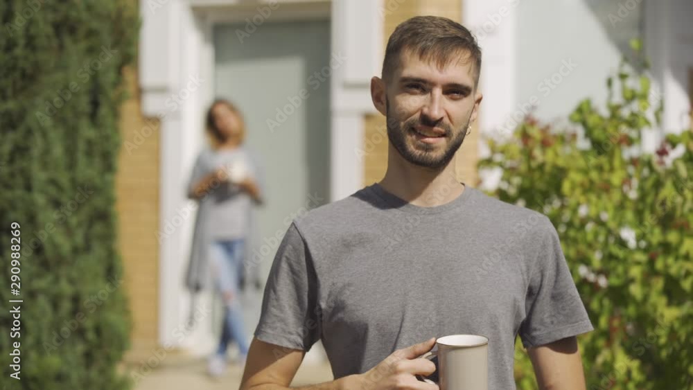 Handsome man standing with the cup of tea in the foreground looking at camera, girl standing on the background. Young family together at the house. Summertime. Handsome man standing with the cup of tea in the foreground looking at camera, girl standing on the background. Young family together at the house. Summertime.