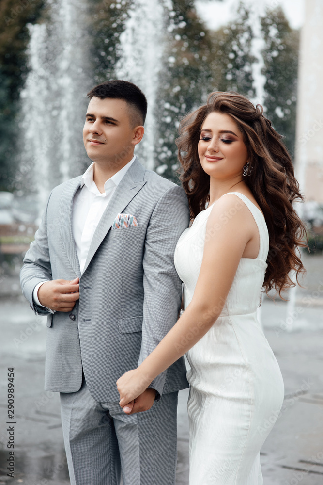 Beautiful long-haired girl in summer dress with her handsome husband in grey suit walking near fountain. Summer date. Happy couple walking near the beautiful fountain in the hot summer day. Beautiful long-haired girl in summer dress with her handsome husband in grey suit walking near fountain. Summer date. Happy couple walking near the beautiful fountain in the hot summer day.