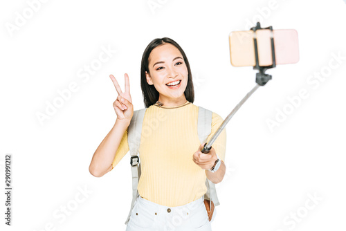 attractive asian woman showing victory sign while taking selfie on smartphone with selfie stick isolated on white