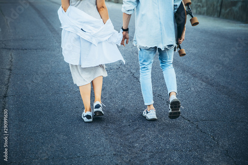 Tanned young caucasian couple, modern lovestory in film grain effect and vintage style. Sunset. Walking on the city's street with longboard, summer evening. Honeymoon concept. Toned in teal orange.