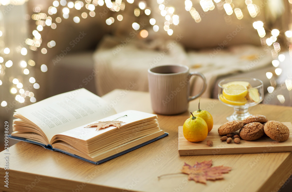 Fototapeta premium hygge and cozy home concept - book, autumn leaves, cup of tea with lemon, almond nuts and oatmeal cookies on table