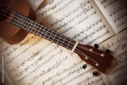 A small four-stringed ukulele with nylon strings lies on old white scattered pages of sheet music.